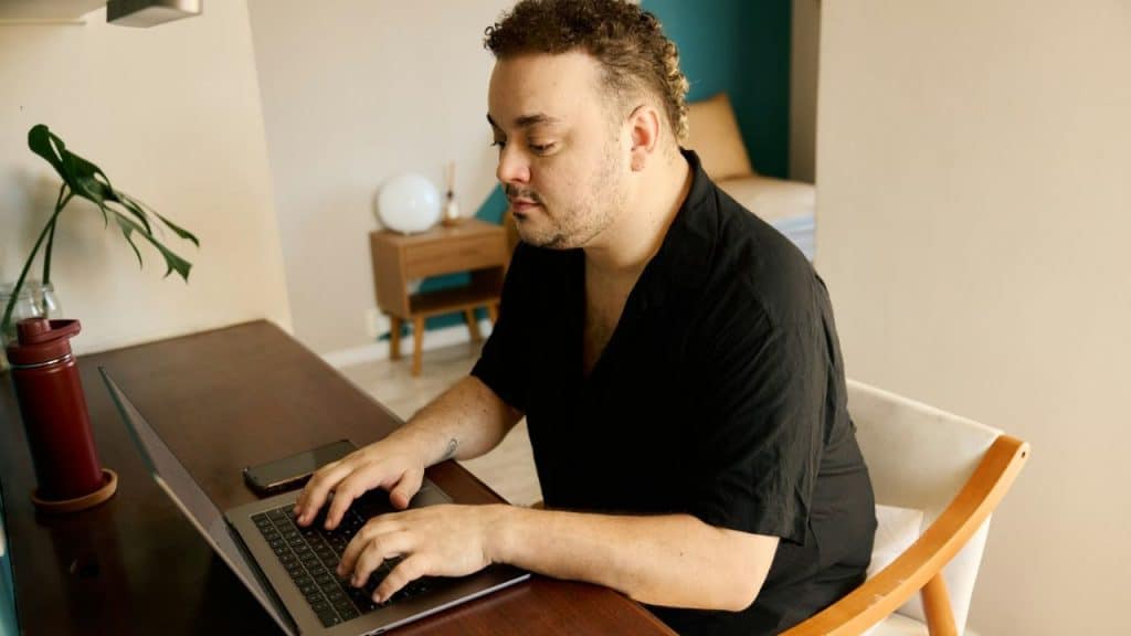 A man with short curly hair and a beard, wearing a black shirt, types on a laptop at a desk.