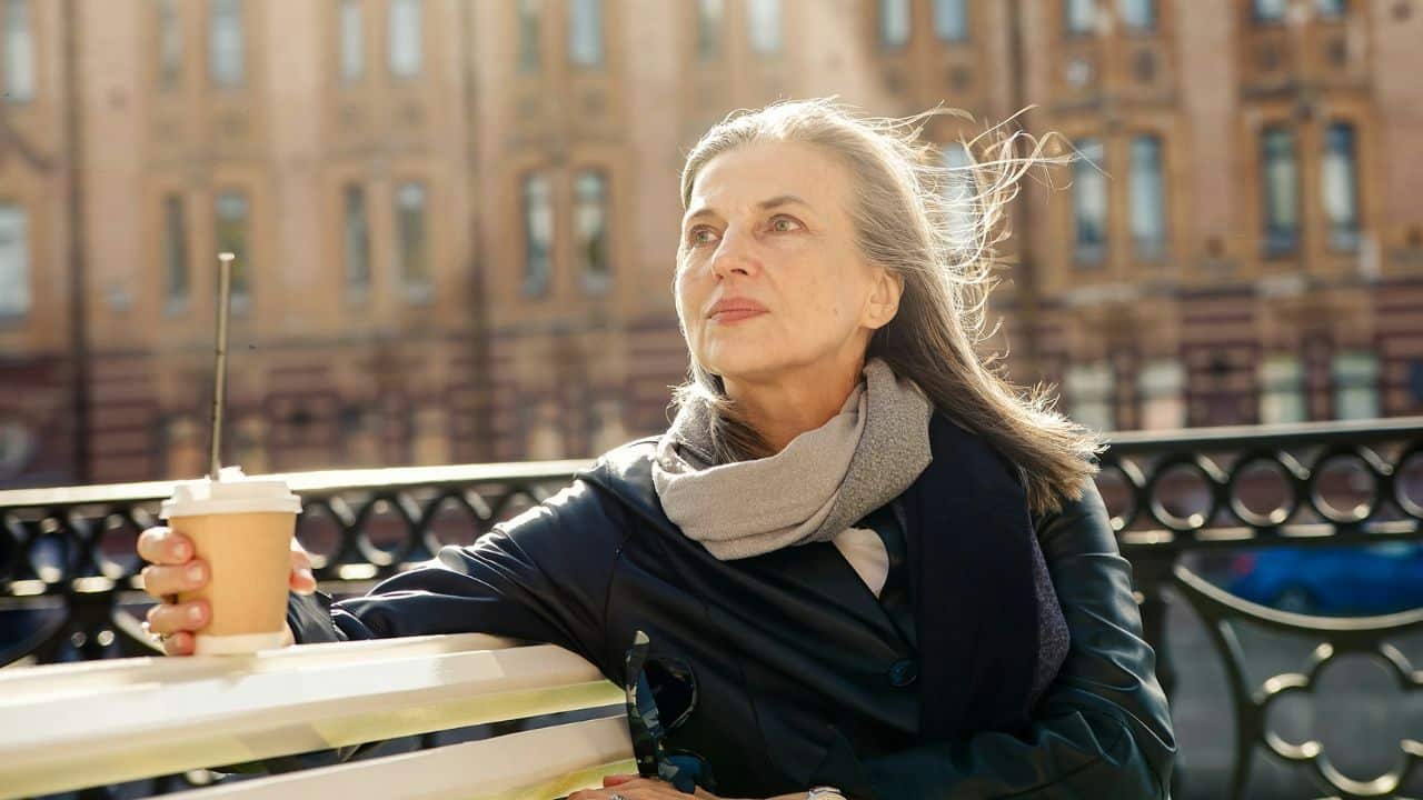 A pensive older woman with long gray hair sits on a bench, holding a cup.