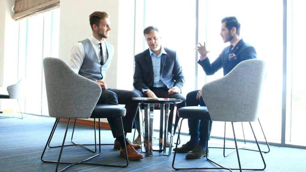 A diverse group of three businessmen converse in a modern, sunlit office lounge.