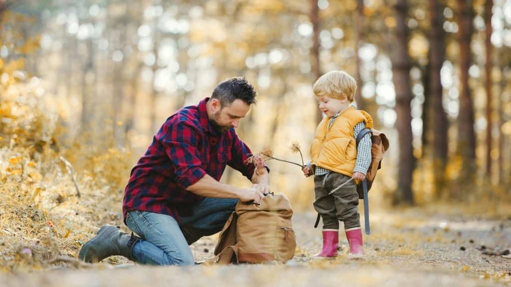A man kneeling by a backpack as a child holds branches in a forest.