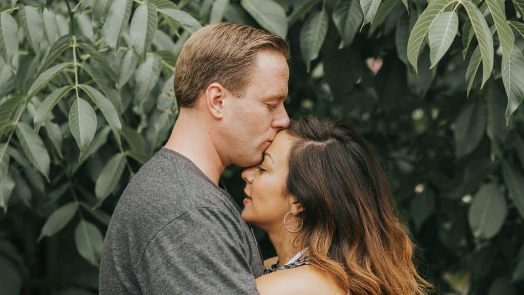 A man gently kisses a woman's forehead, both smiling softly, surrounded by greenery.