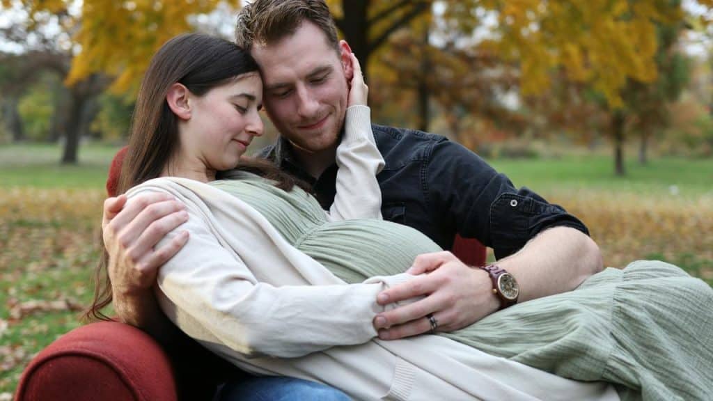 A pregnant woman cradling her belly in her partner’s embrace outdoors.