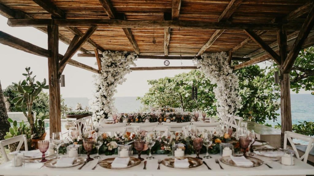 An outdoor wedding reception table set beneath a flower-draped wooden pavilion by the water.