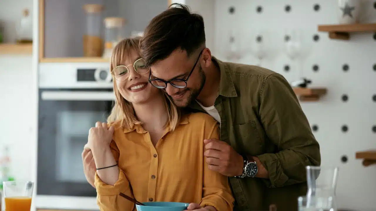 A man embraces a smiling woman in glasses, holding a bowl.