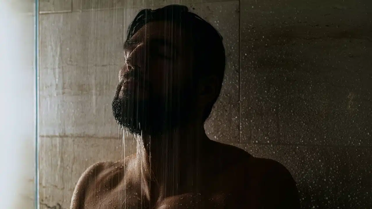 A man with a beard stands under a shower, water running down his face.