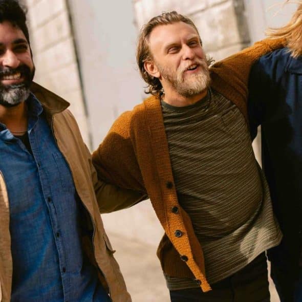 A trio of casually dressed men walking arm in arm down a sunlit street, smiling and laughing.