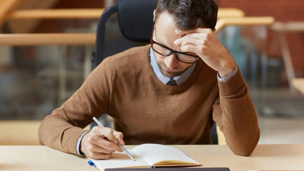 A man in a brown sweater and glasses writes in a notebook, resting his head on his hand.