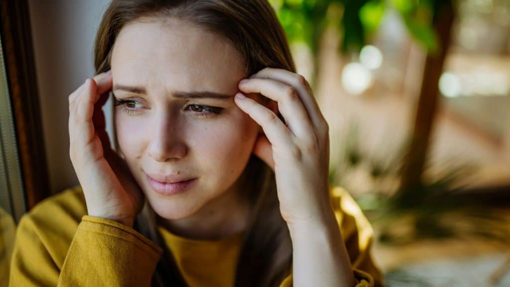 A distraught young woman with hands on her head looks out a window.
