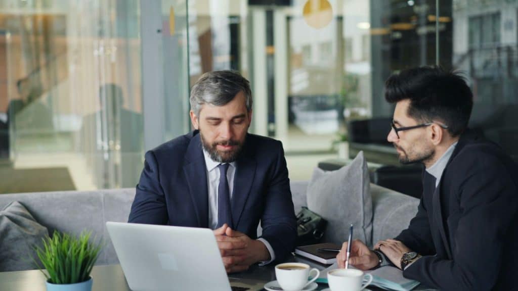 A bearded man in a suit looks down at a laptop while another man in glasses talks.