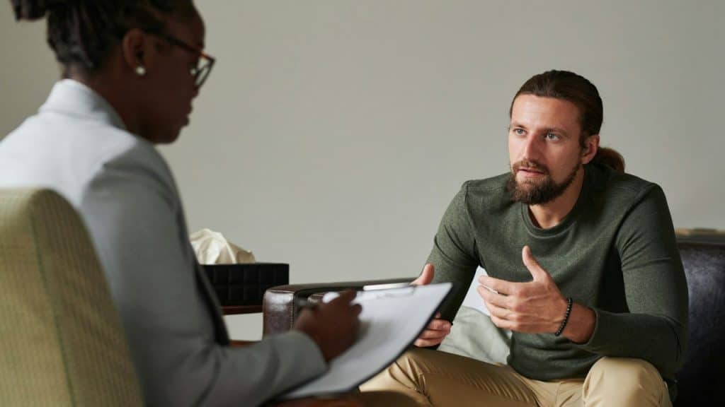 A male patient gestures while speaking to a female therapist taking notes.