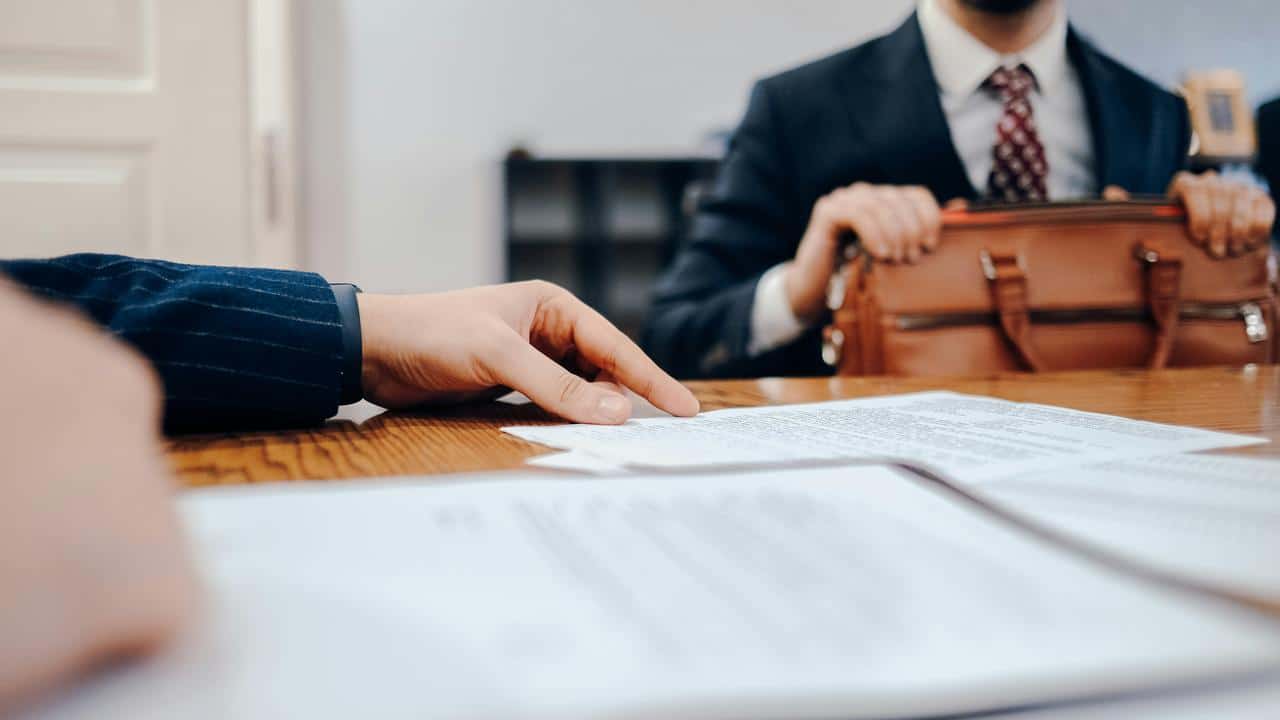 A person pointing at documents on a desk while another holds a briefcase.