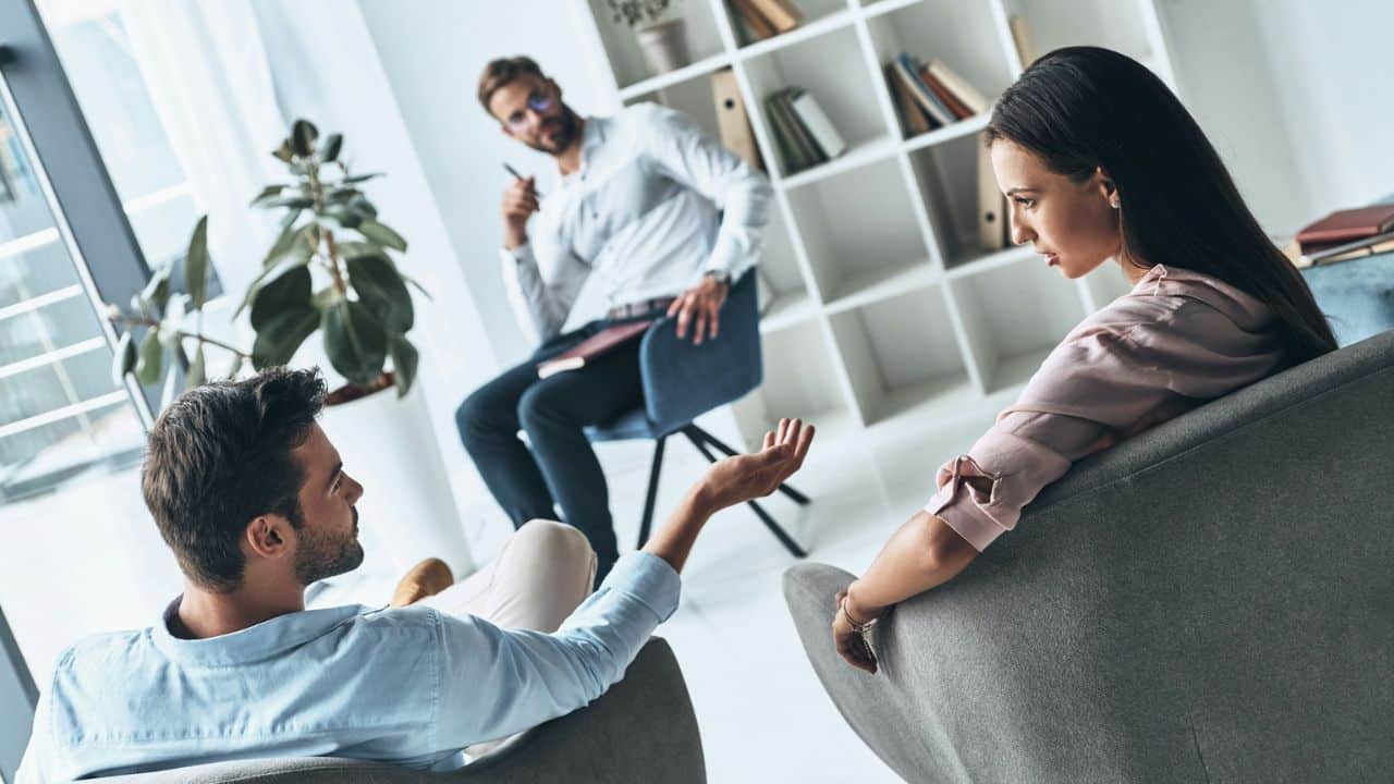 A man gestures towards a woman as a therapist listens in the background.