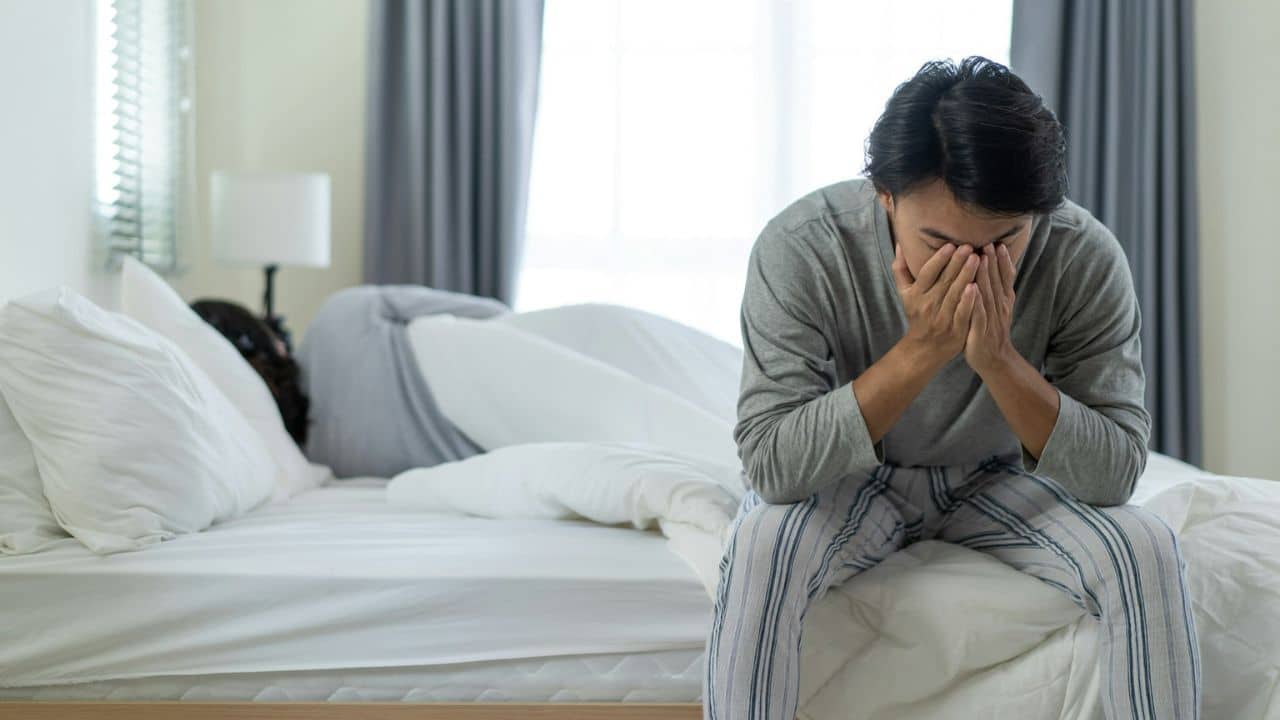 A man sits on a bed with his face in his hands, while a person is lying in the bed behind him.