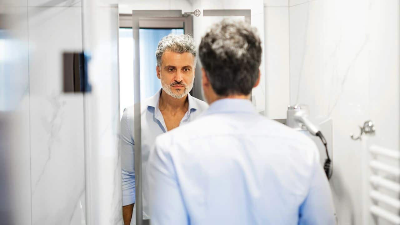 A man with graying hair looks at his reflection in a bathroom mirror.