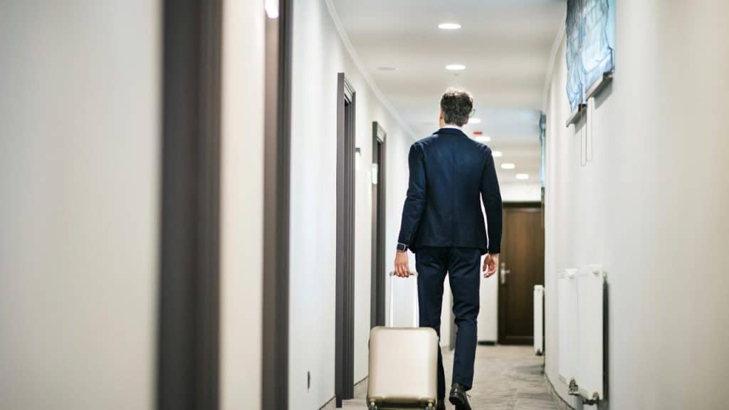A businessman walking down a hotel hallway pulling a rolling suitcase.