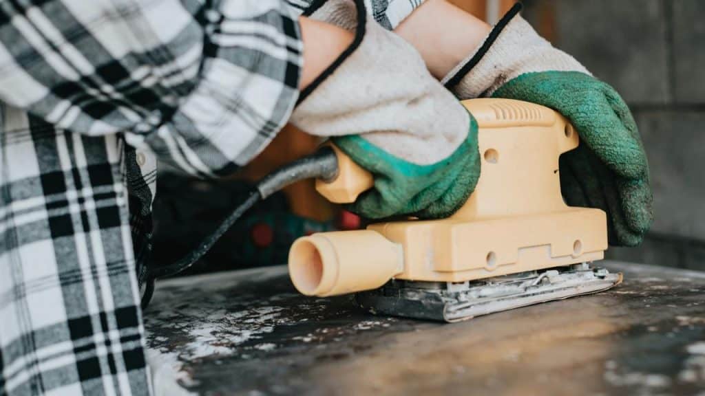 A green‑gloved hand operating a yellow electric sander on a work surface.