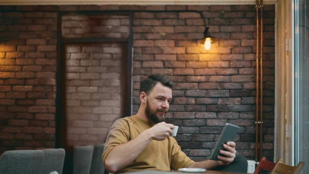 A bearded man sipping coffee while reading a tablet in a brick‑walled café.