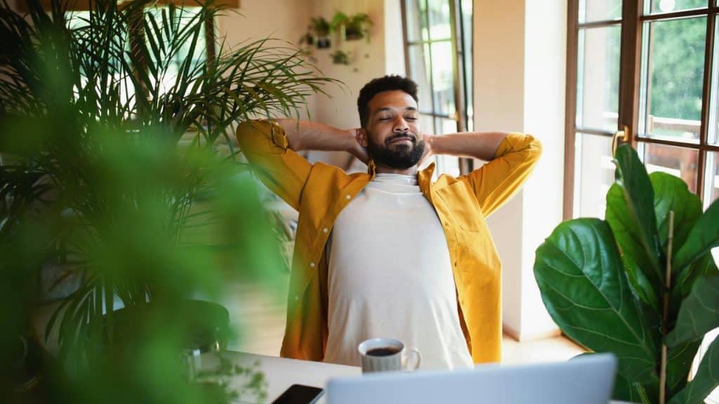 A man leaning back at his desk with his eyes closed amid green plants.