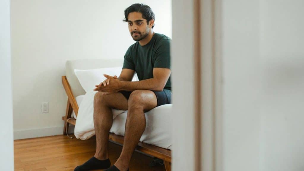 A thoughtful man with dark hair and a beard sits on a bed, hands clasped.