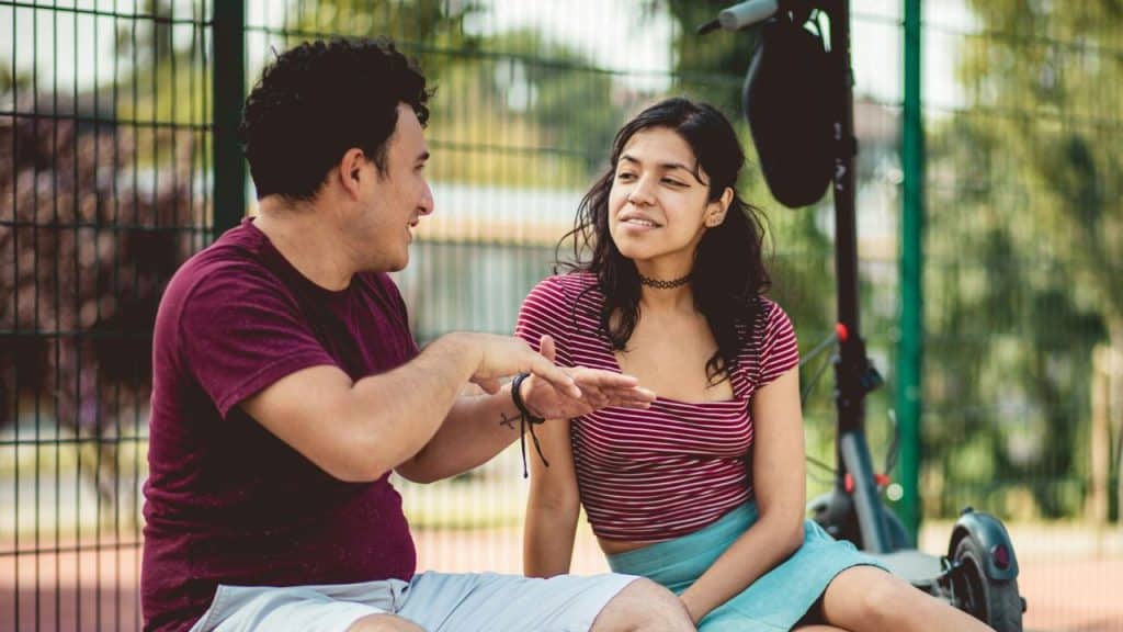 A man gestures while talking to a woman sitting next to him outdoors.