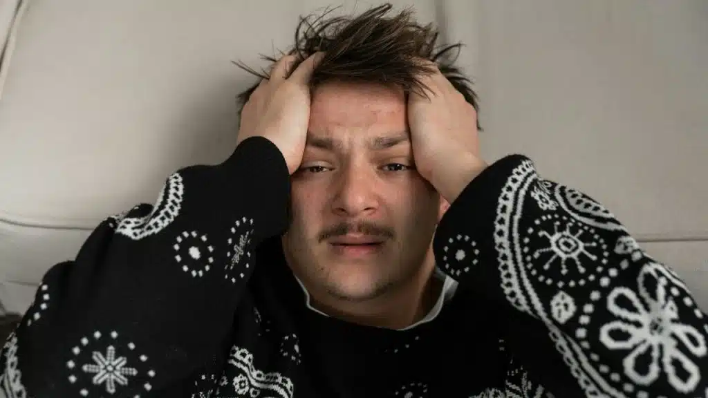 A person with disheveled hair and a mustache holds their head, looking stressed.