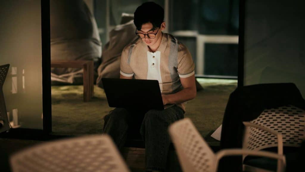 A man wearing glasses and a polo shirt works on a laptop in a dimly lit room.
