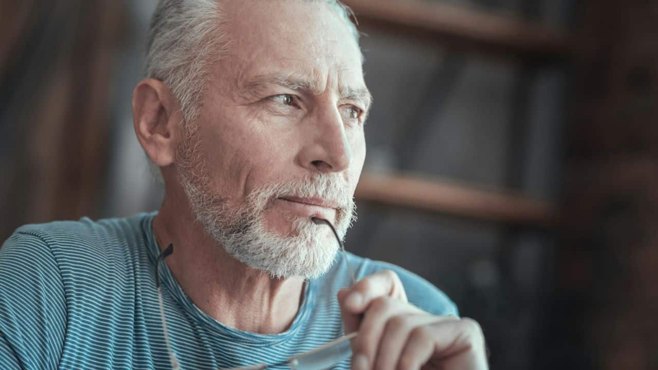 A contemplative, grey-bearded man in a striped shirt holds glasses to his mouth.