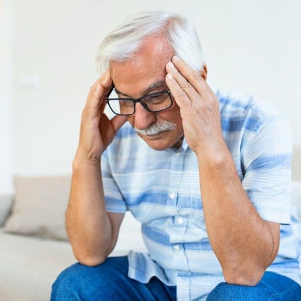 A grey-haired man with glasses and a mustache sits on a couch, holding his head.