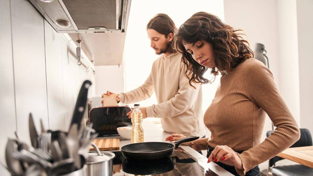 A woman cooks on a stovetop while a man makes toast in the background.