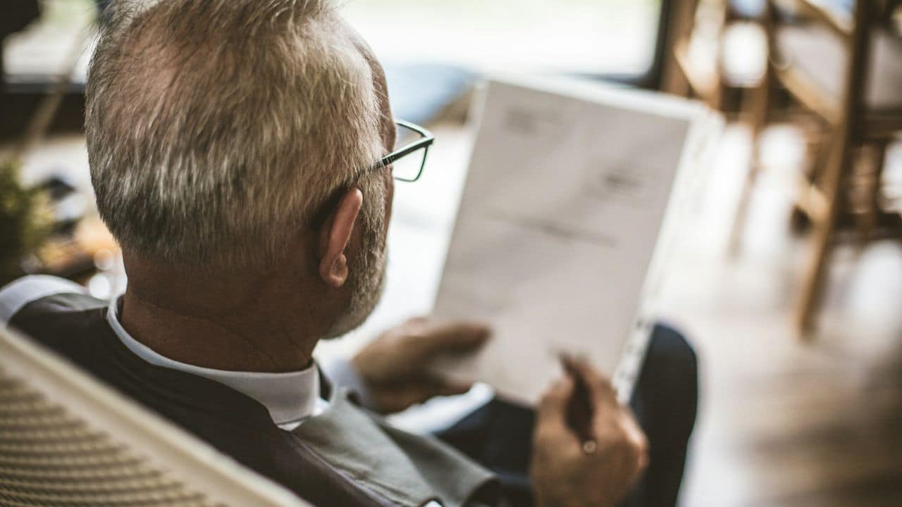 A grey-haired man with glasses, viewed from behind, reads a document.