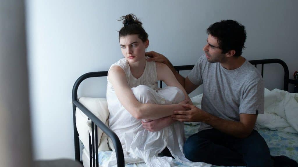 A man in a grey shirt comforts a woman in a white dress sitting on a bed.