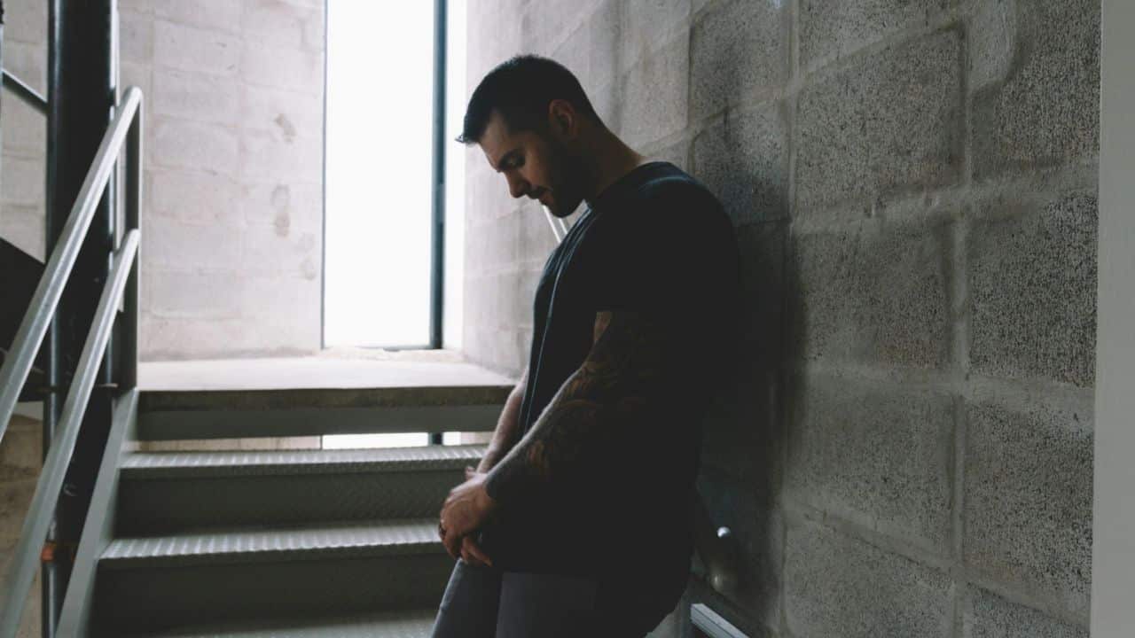 A man with a beard and tattoos stands leaning against a concrete wall, looking down.