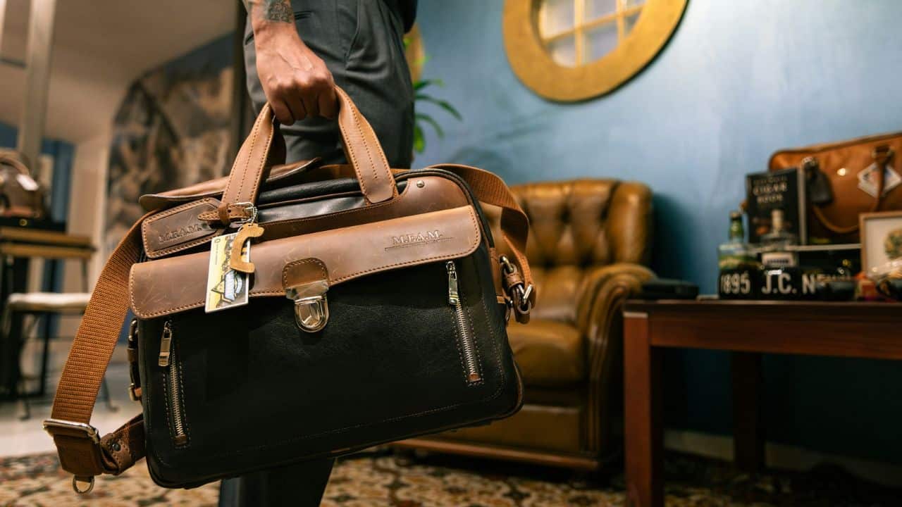 A man in a suit holds a brown and black leather messenger bag, entering a room.