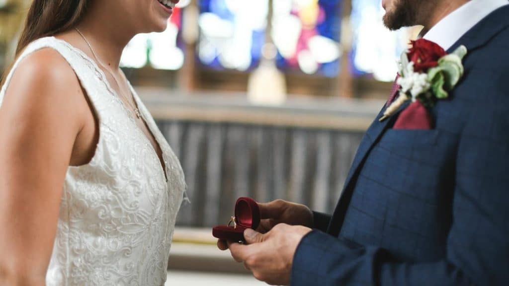 A groom in a blue suit offering a ring in a red velvet box to a bride in a white dress.