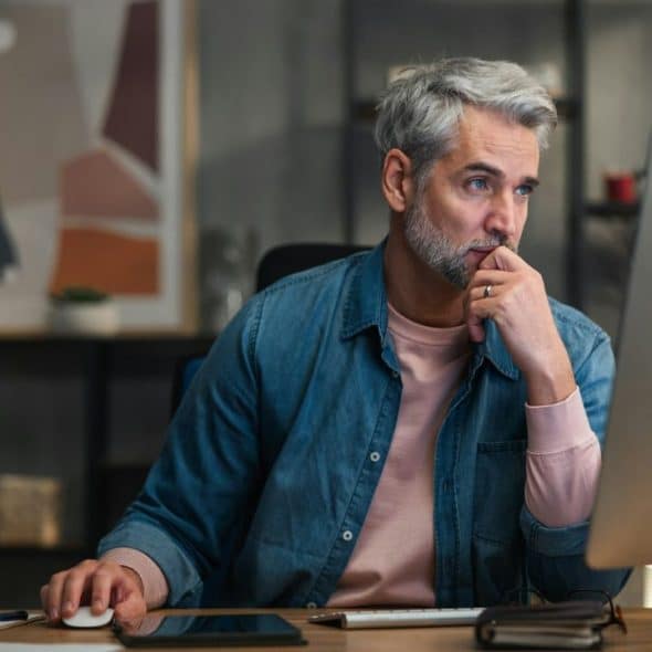 A focused man with gray hair and a beard uses a computer at a desk.