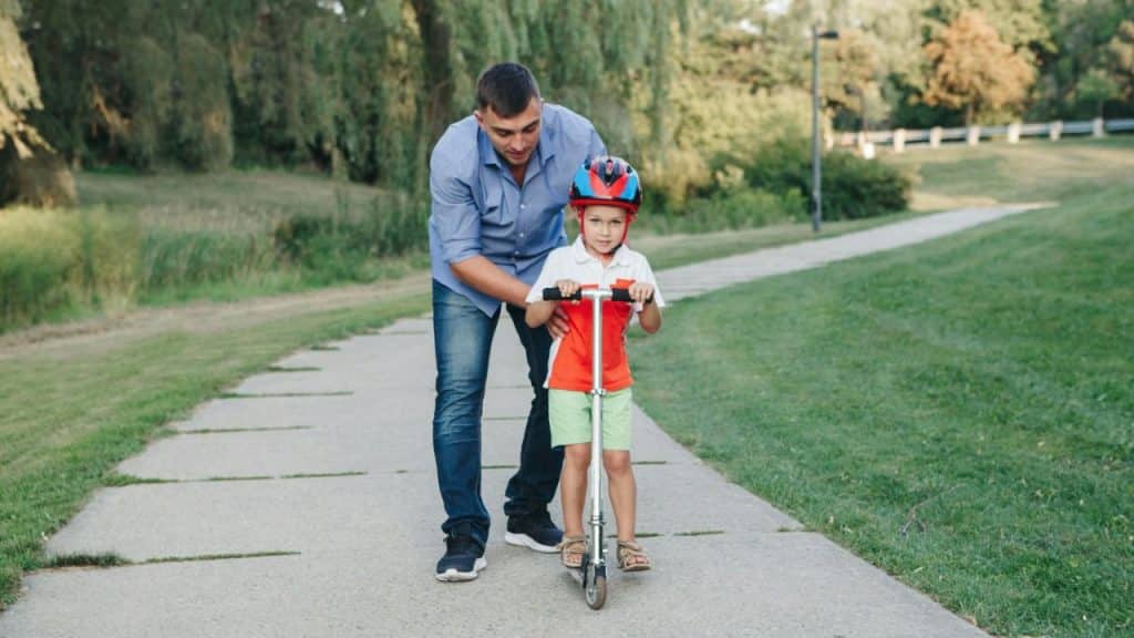 A man helps a young boy in a helmet and red shirt ride a scooter on a paved path.