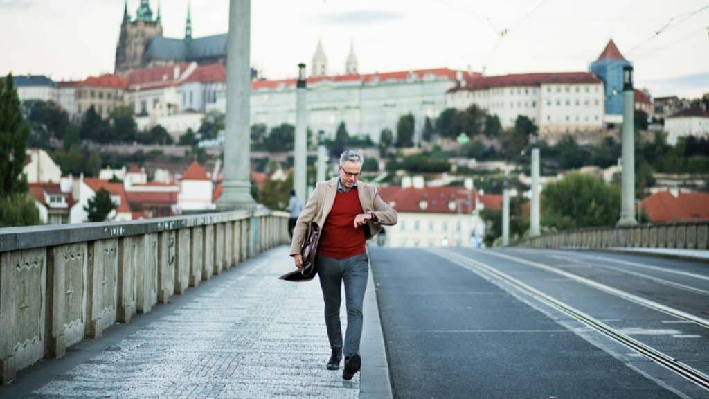 A man in a brown jacket and red sweater walks on a bridge, looking at his watch, with a city in the background.