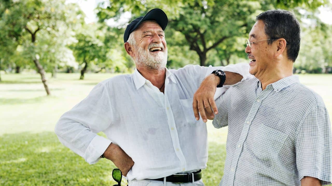 A smiling older man in a cap leans on another man in a park.