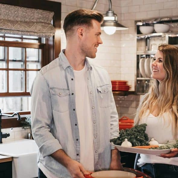 A couple in a kitchen holding fresh vegetables and cookware.