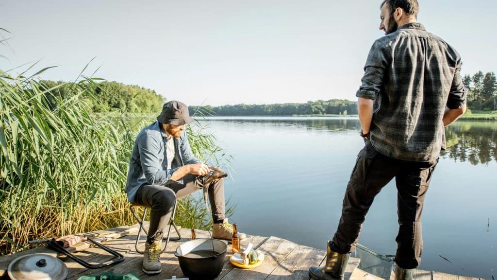 A man cleaning a fish on a lakeside dock as a friend watches.