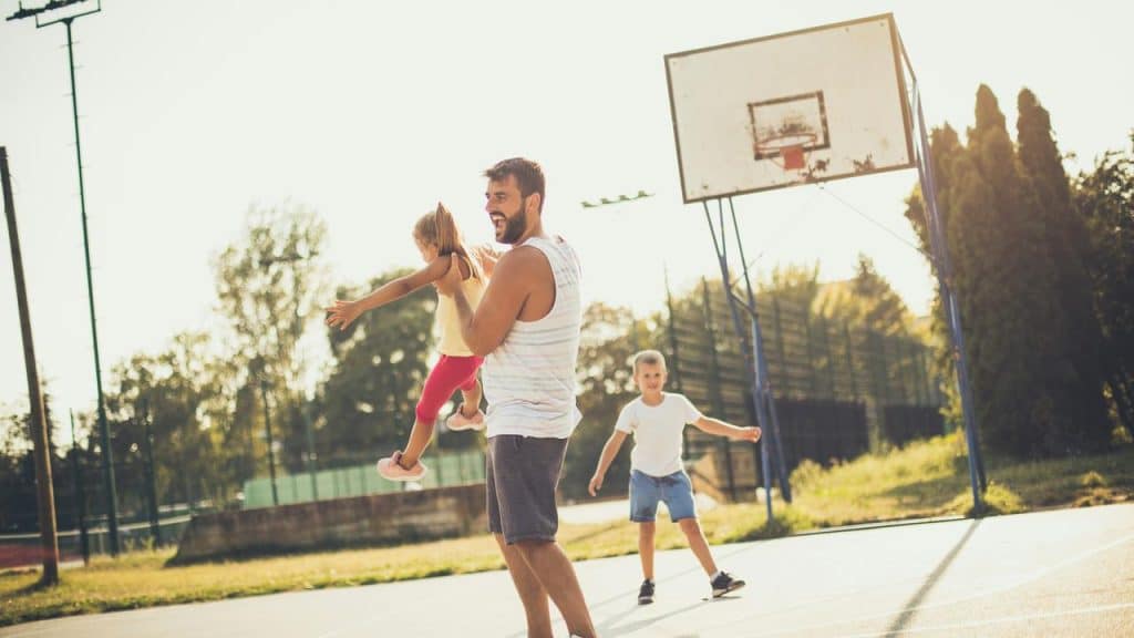 A father swinging his daughter on an outdoor basketball court as his son watches.