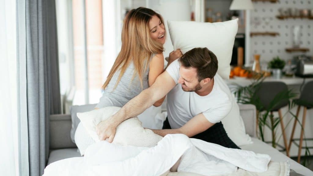A smiling couple is playfully engaged in a pillow fight on a bed.