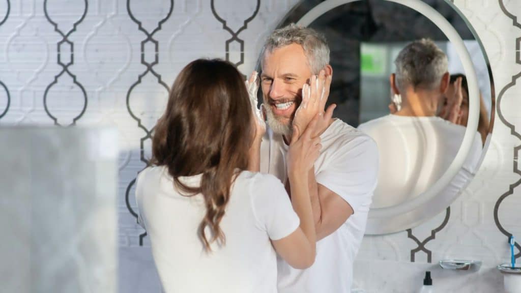 A woman applies cream to a smiling man's face in a bathroom with a mirror.