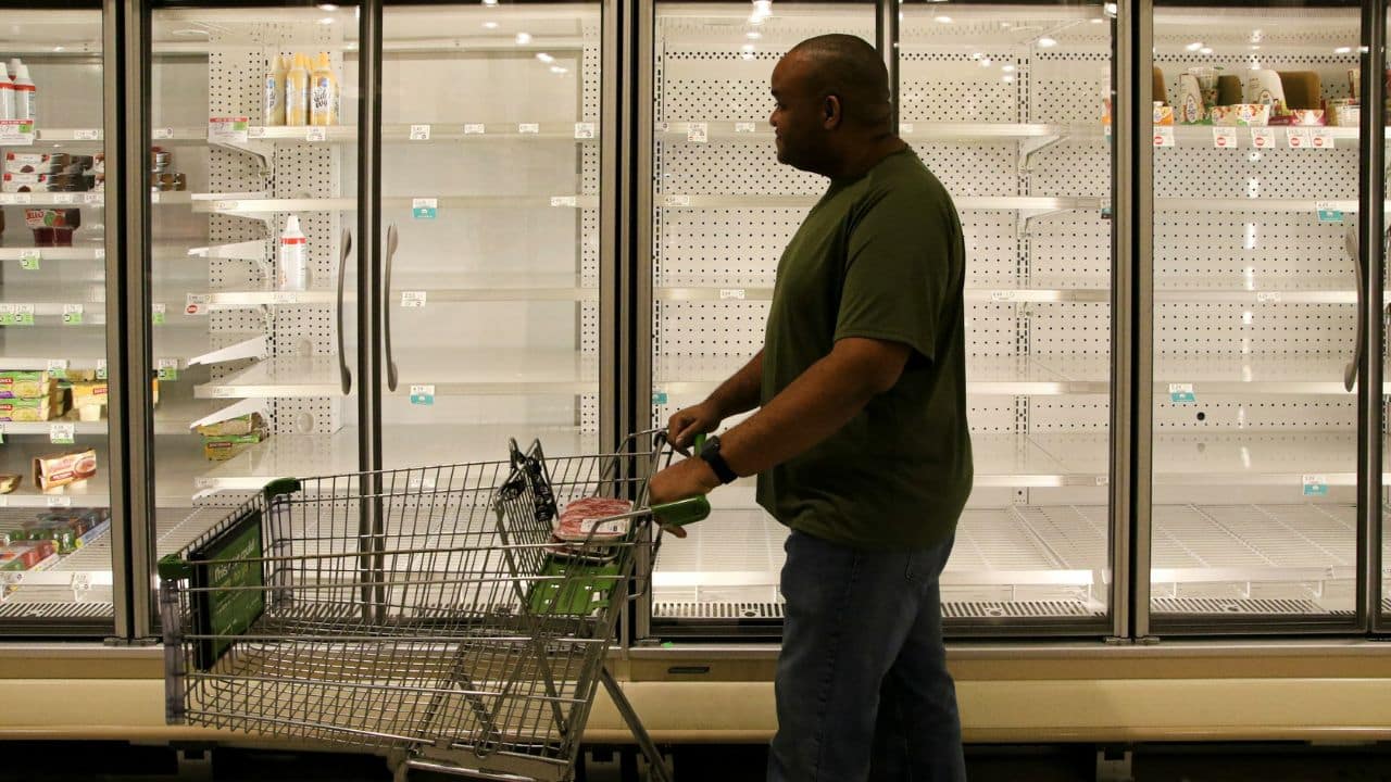 A man pushes an empty shopping cart past mostly bare grocery store freezer shelves.