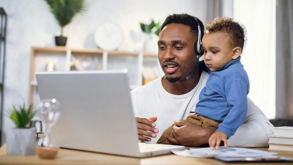 A man with a headset holding a toddler while using a laptop.