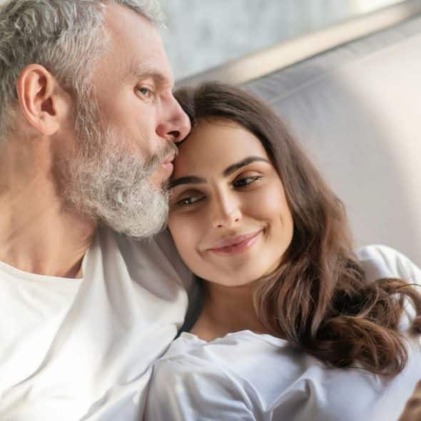 A gray-haired man kisses a woman with dark hair who holds a cup.