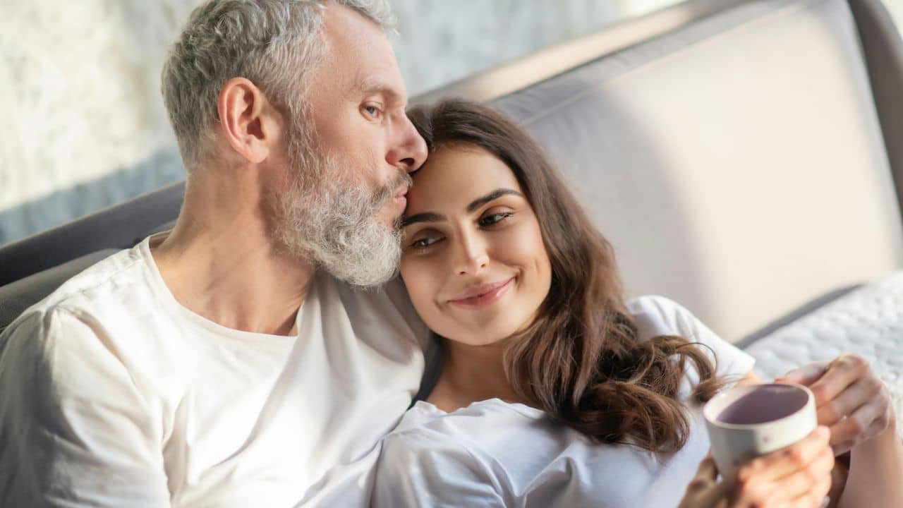 A gray-haired man kisses a woman with dark hair who holds a cup.