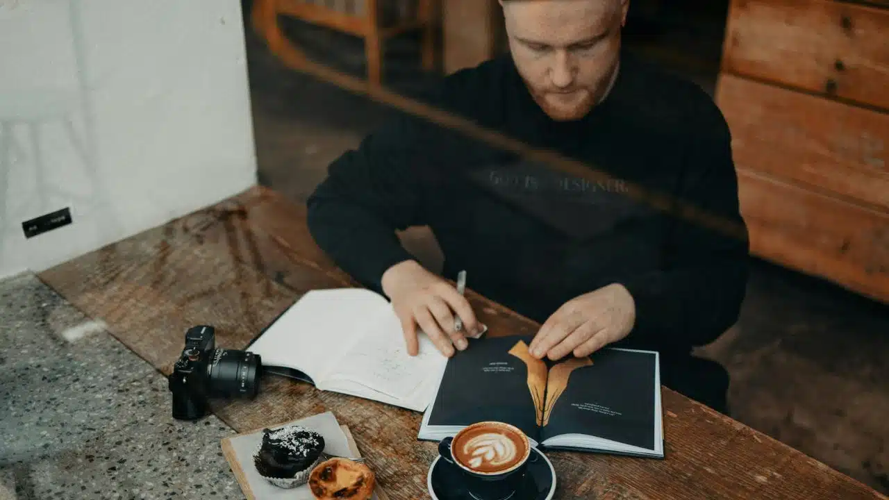 A man in a black sweatshirt reads a book at a wooden table with coffee and pastries.