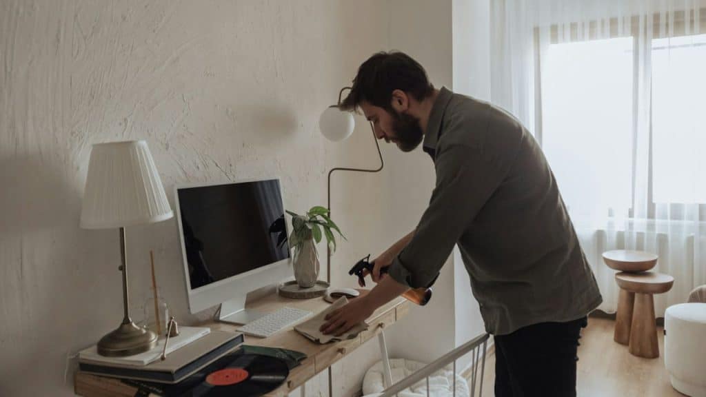 A man with a beard wipes down a desk next to a computer.