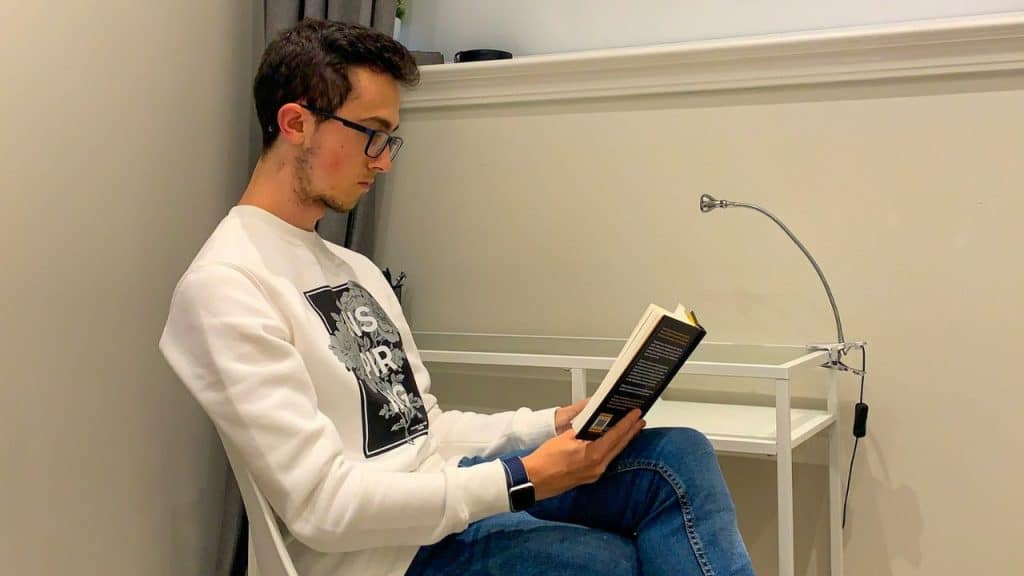 A young man in glasses and a white sweatshirt sits reading a book.